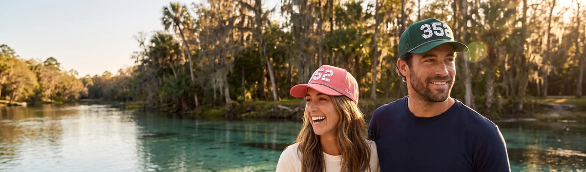 Couple in Palmr 352 Signature hats, pink and green, beside a clear spring run under cypress in Ocala springs country