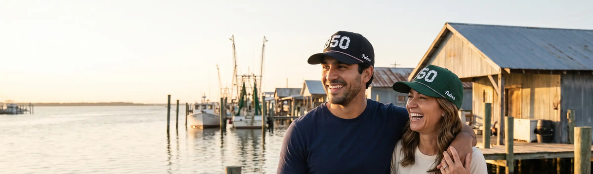 Couple in Palmr 850 Signature hats on a dock with shrimp boats and tin-roof fish houses in the Florida Panhandle