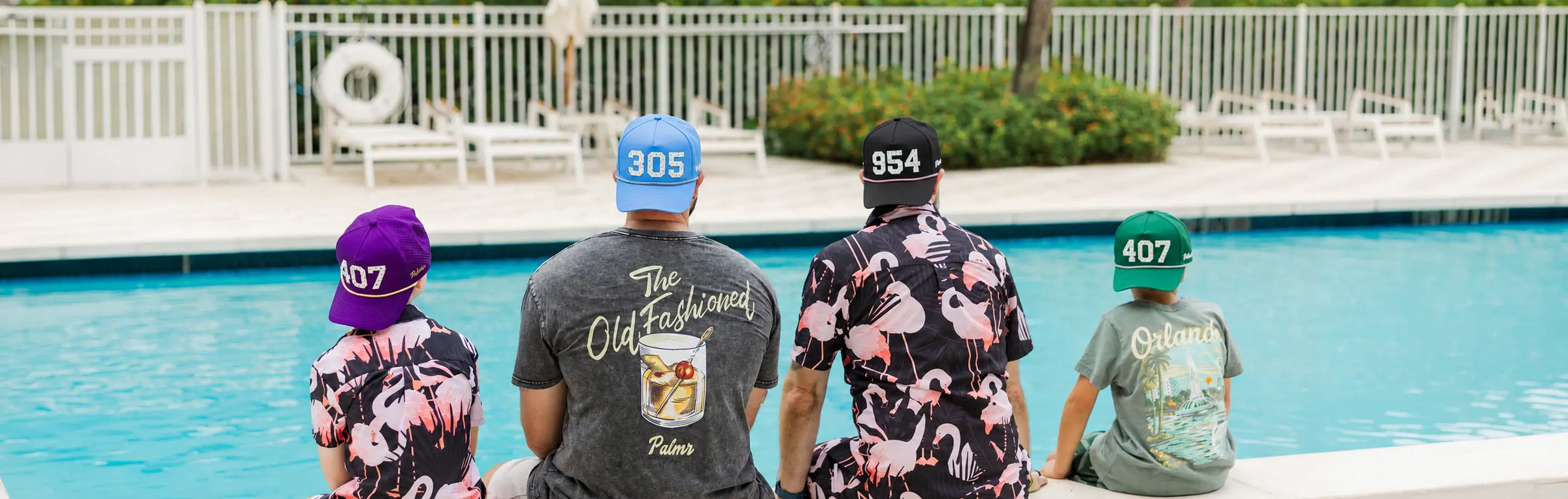 Four people sitting on the edge of a swimming pool, wearing matching pink flamingo-print shirts and colorful caps. The shirts feature the text 'The Old Fashioned' and 'Orlando' on the back. 407 305 954 hats
