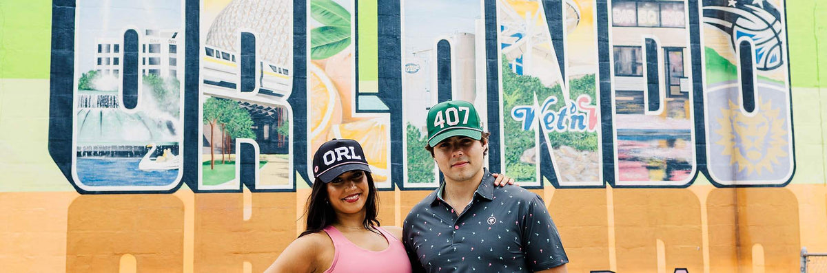 A couple in Palmr ORL and 407 rope hats in front of the Orlando mural