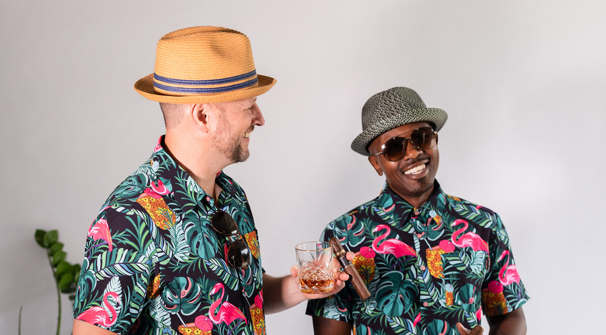 Two men in matching Palmr button-up shirts with a flamingo, palm and pineapple print, one in a straw fedora with a drink