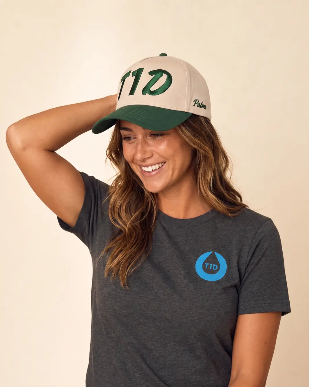 A woman wearing a dark gray t-shirt with a blue T1D logo and a beige baseball cap with a green brim and green T1D lettering, posing against a plain beige background.