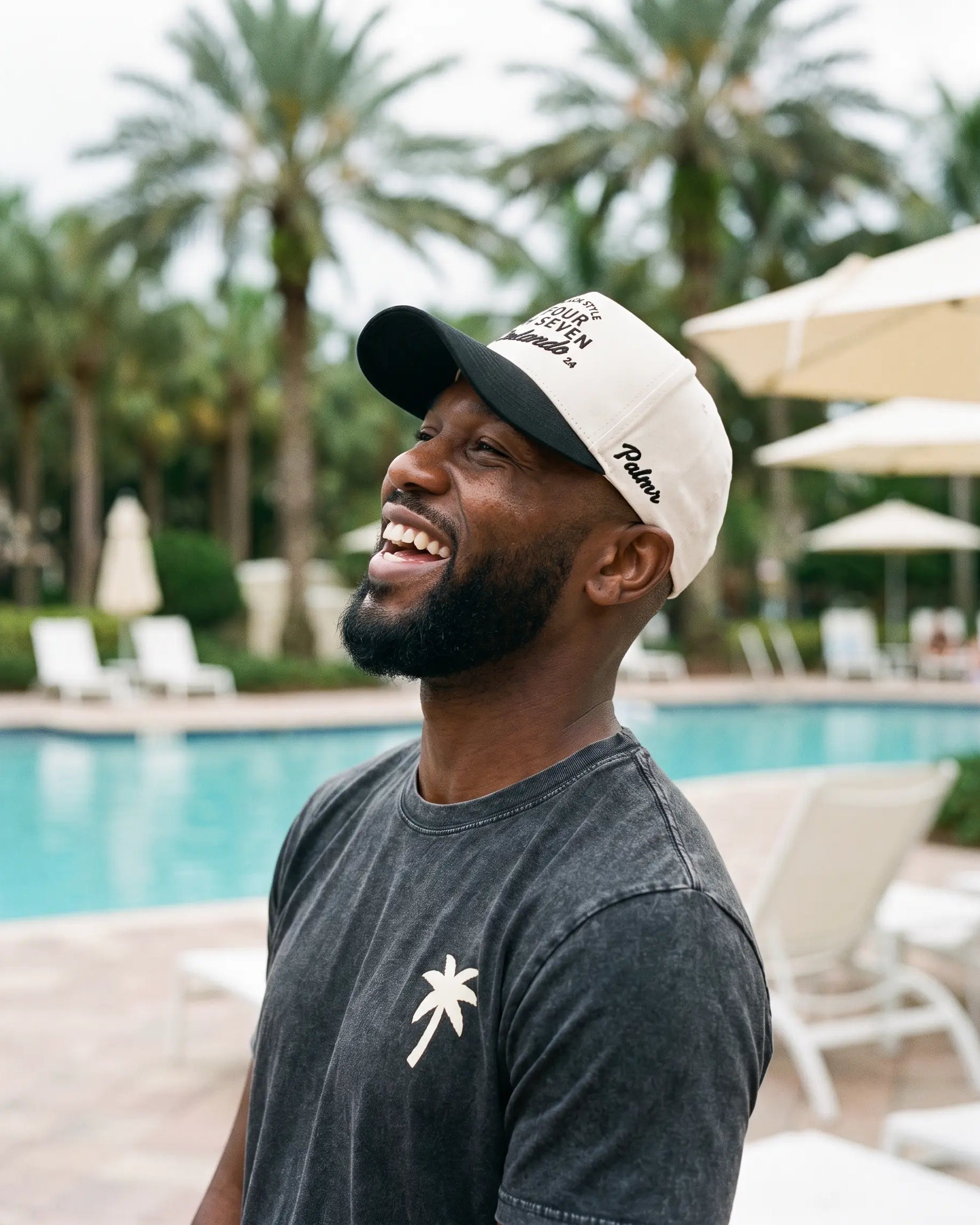 A man smiling outdoors by a swimming pool, wearing a black and off-white baseball cap with 'Palm Beach' and 'The Palm Club' text, and a dark gray t-shirt with a white palm tree graphic.