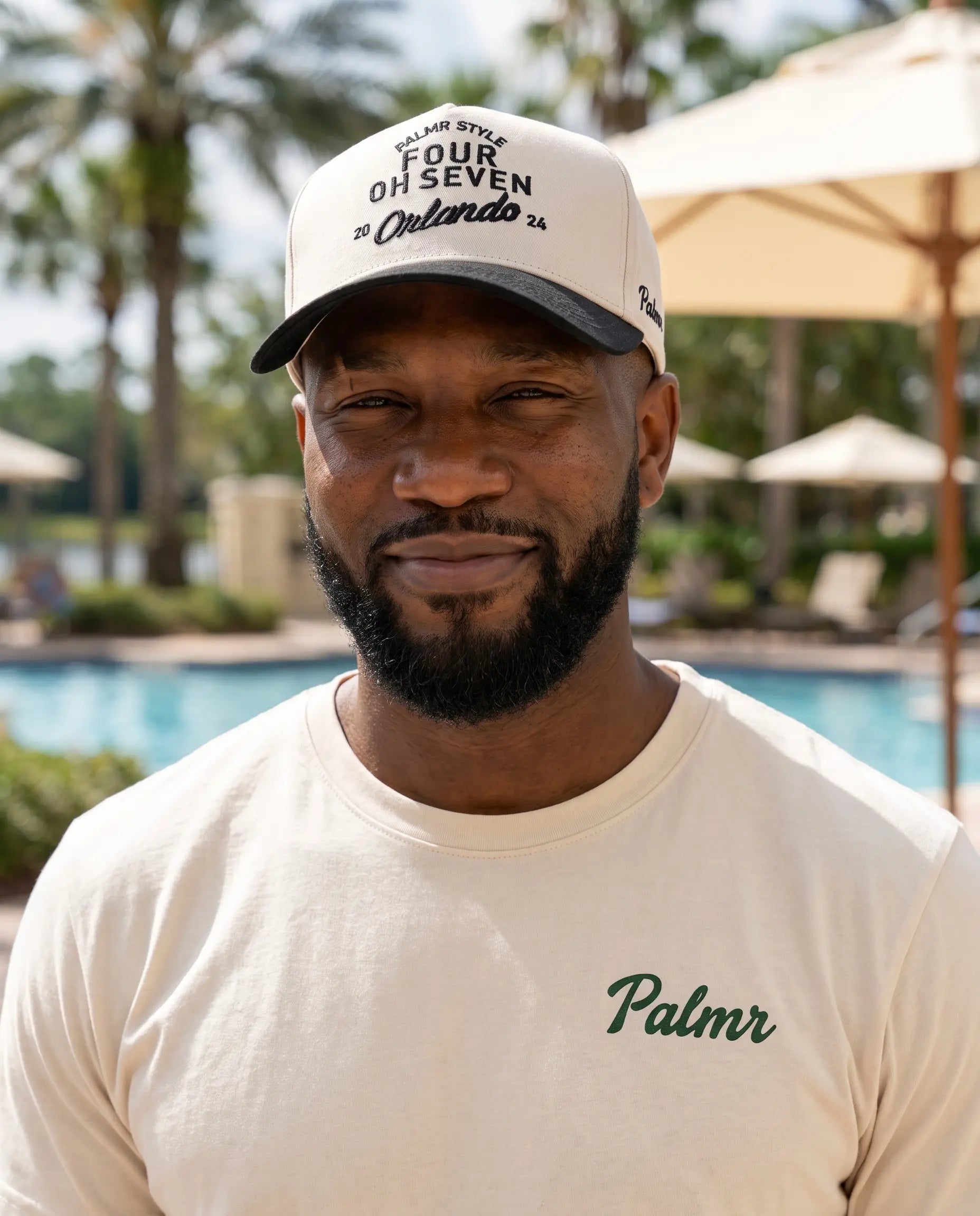A man wearing a cream-colored t-shirt with 'Palmr' printed in green on the chest and a matching cream trucker hat, standing in front of a blurred resort poolside background.