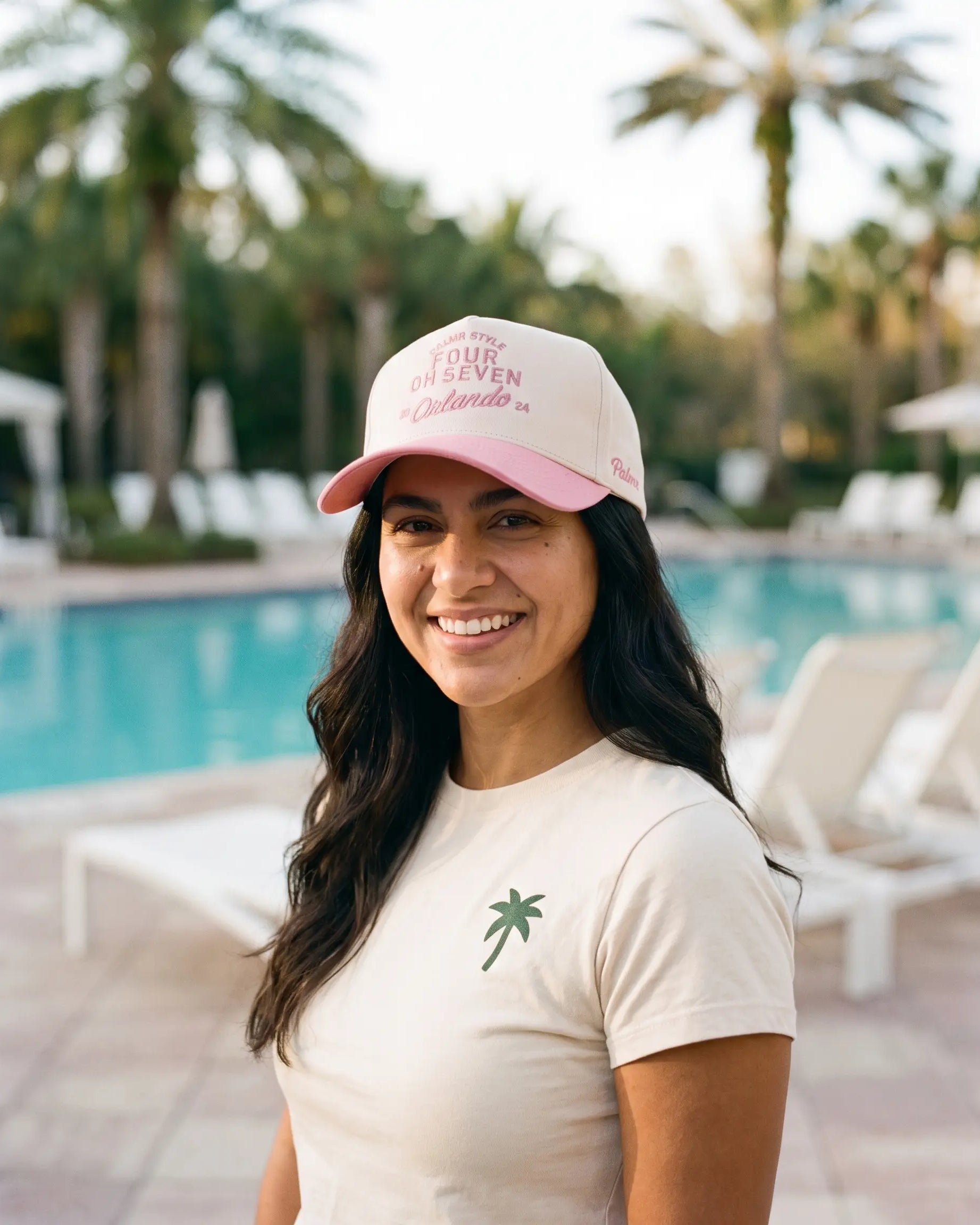 A smiling woman standing outdoors by a swimming pool wearing a light pink and white baseball cap with pink text that reads 'SURF OR DIE SEVEN Orlando' and a cream T-shirt with a small green palm tree graphic.