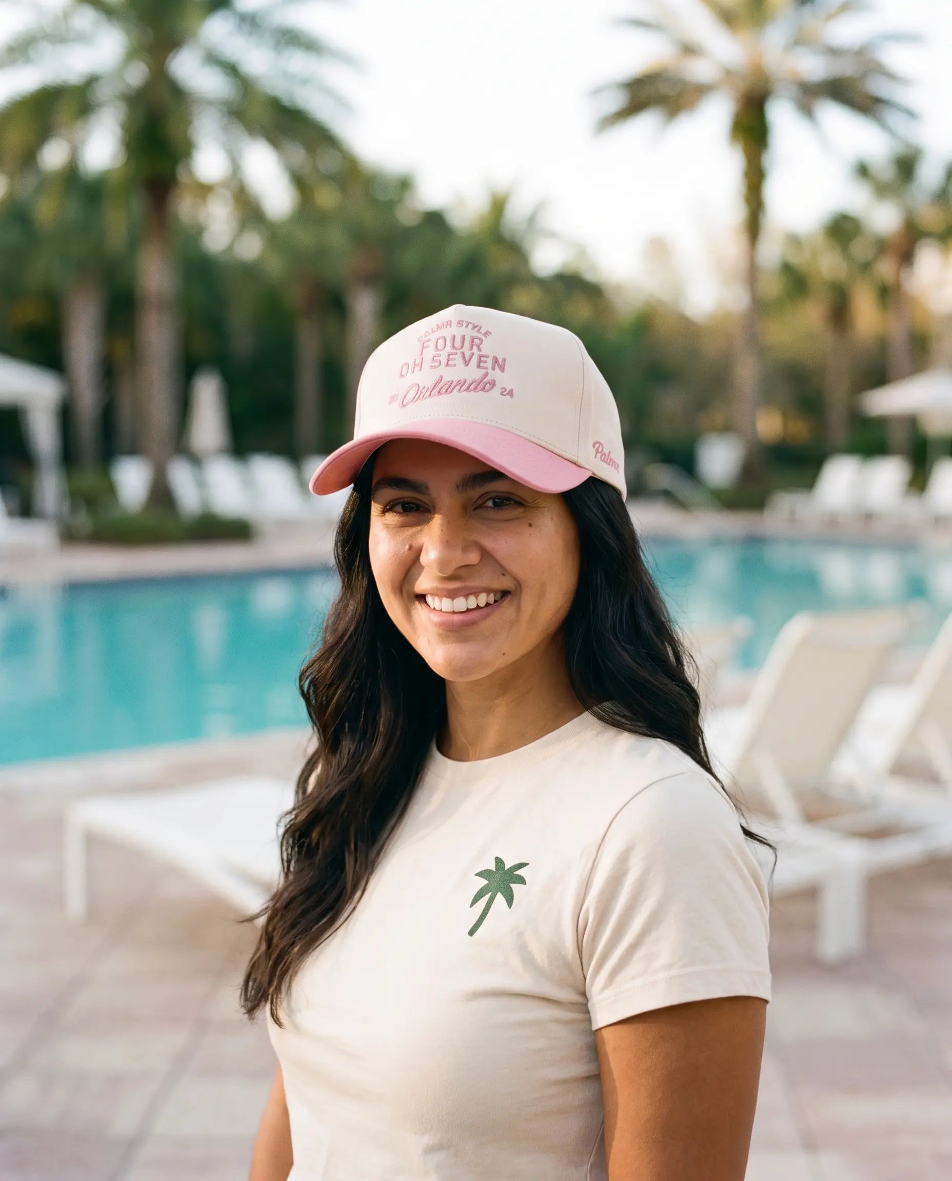 A smiling woman standing outdoors by a swimming pool wearing a light pink and white baseball cap with pink text that reads 'SURF OR DIE SEVEN Orlando' and a cream T-shirt with a small green palm tree graphic.