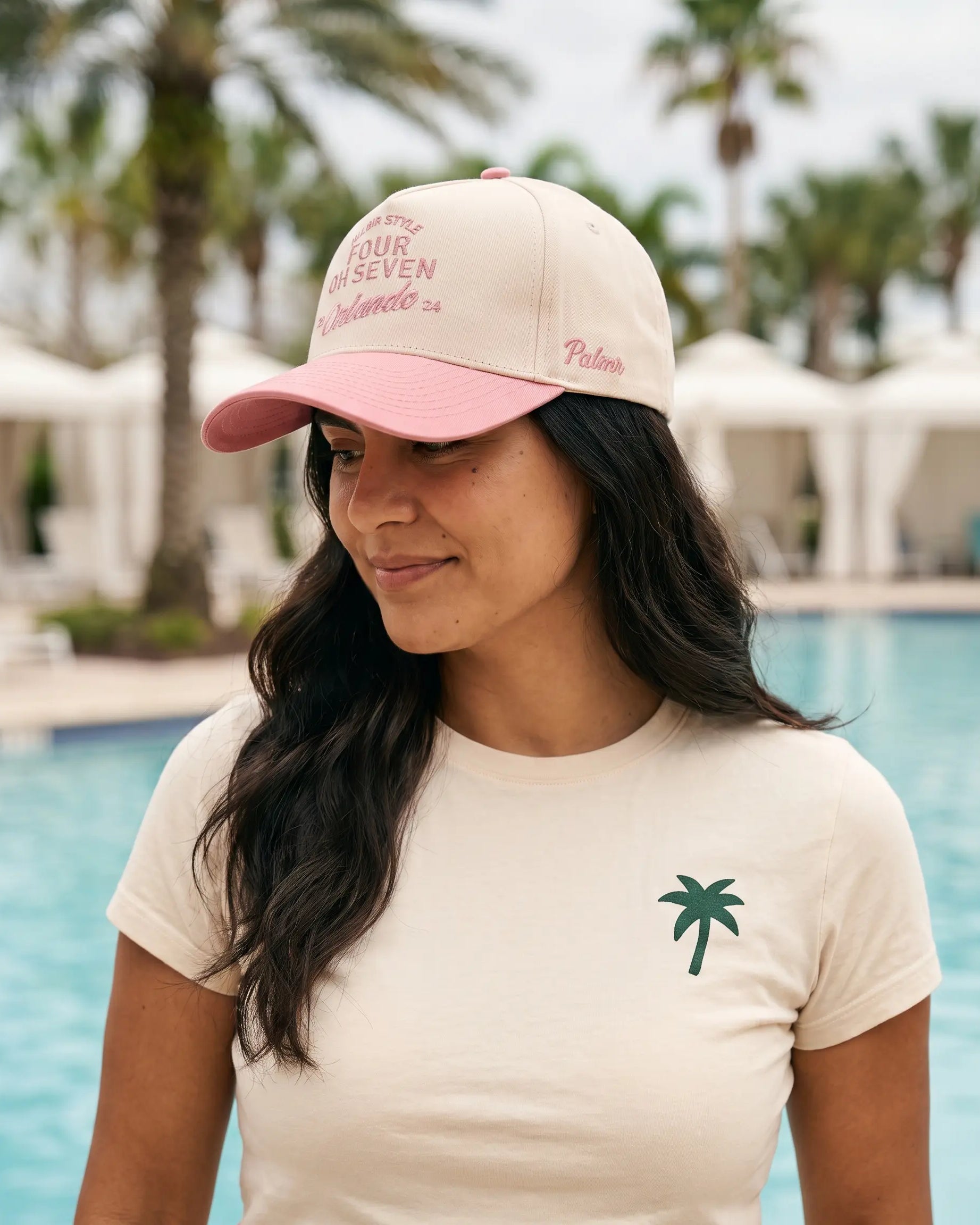 A woman wearing a cream and pink baseball cap with pink embroidered text and a cream t-shirt with a green palm tree logo, standing in front of a resort pool with palm trees in the background.