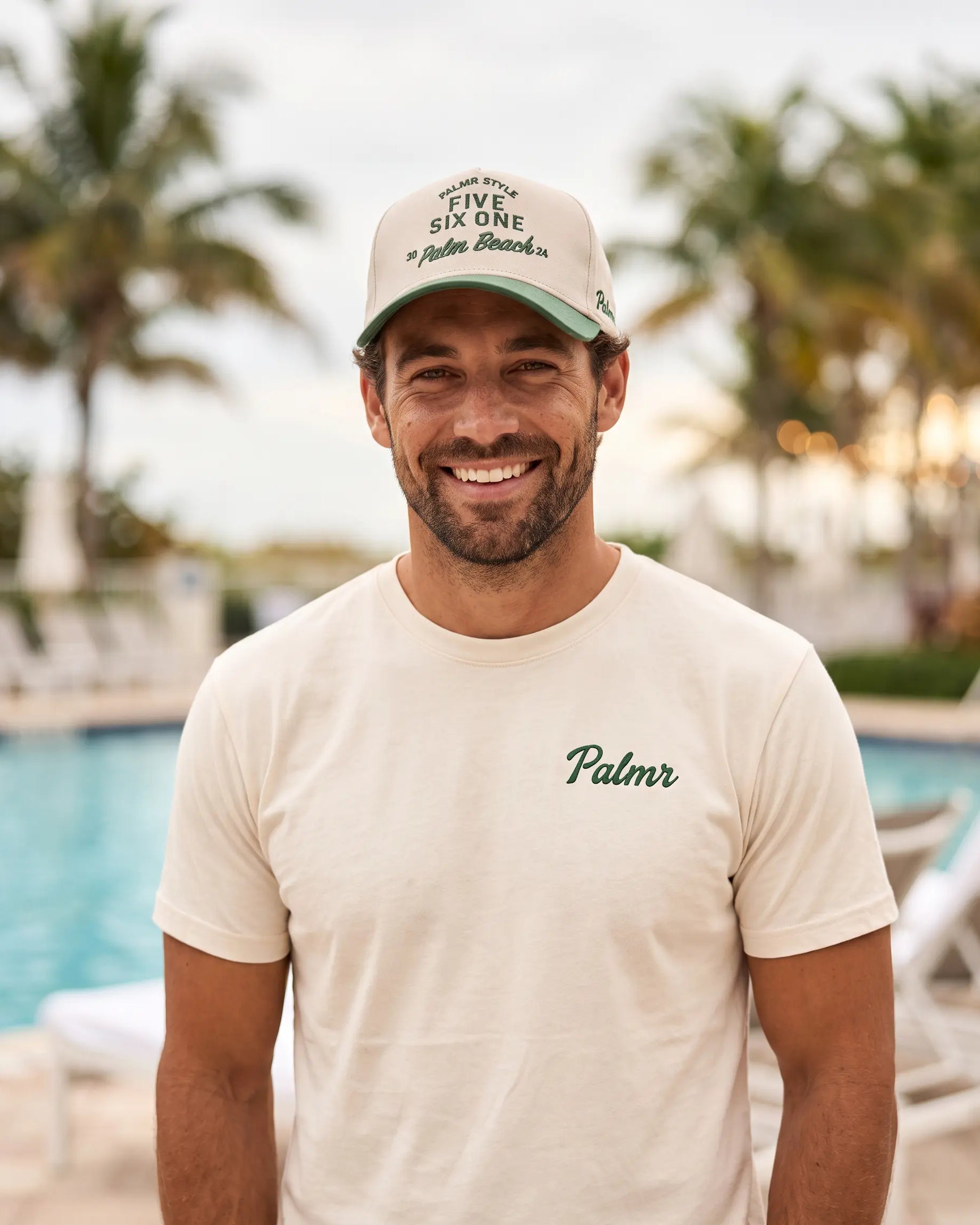 A smiling man wearing a cream-colored t-shirt with 'Palmr' printed on the chest and a matching cream cap with green text, standing in front of a blurred resort pool and palm trees.