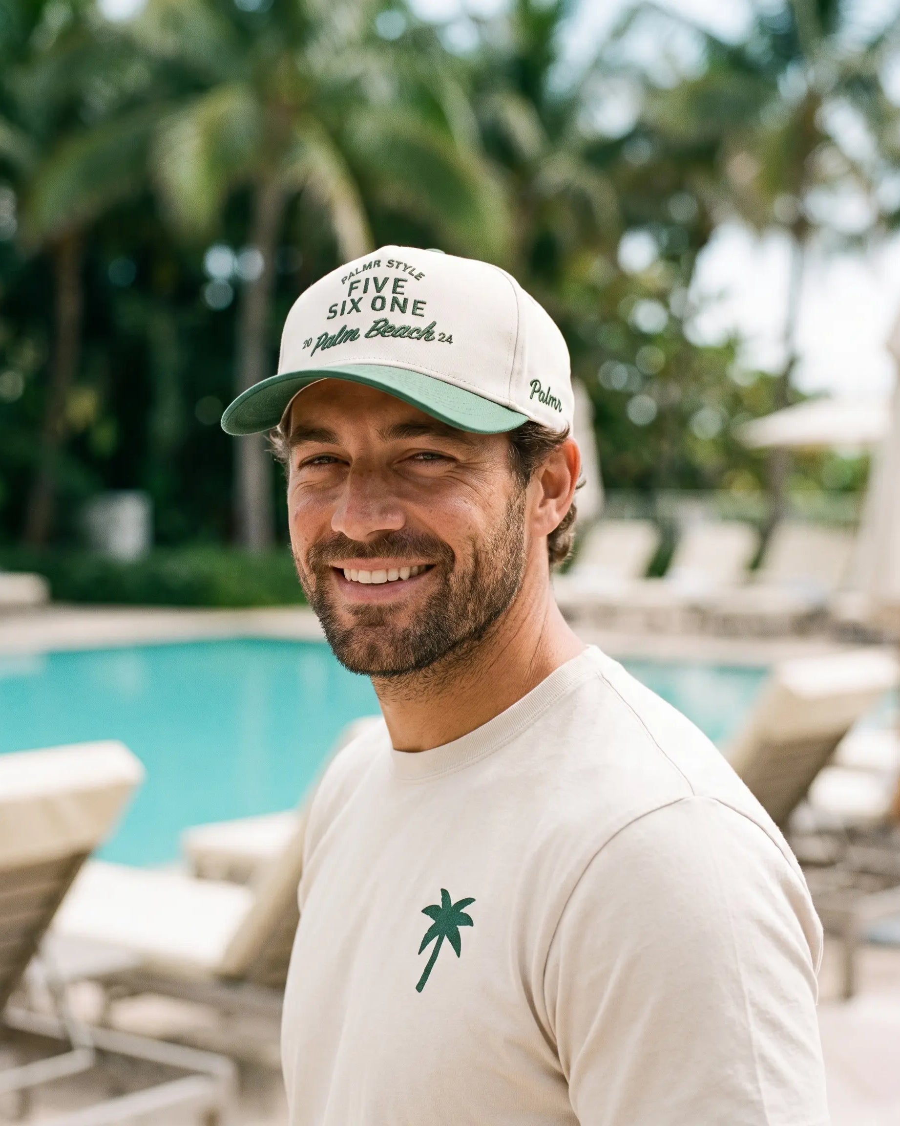 A smiling man wearing a cream-colored baseball cap with green lettering that reads 'FIVE SIX ONE Palm Beach FL' and a matching cream t-shirt with a green palm tree logo, standing in front of a tropical pool with palm trees in the background.