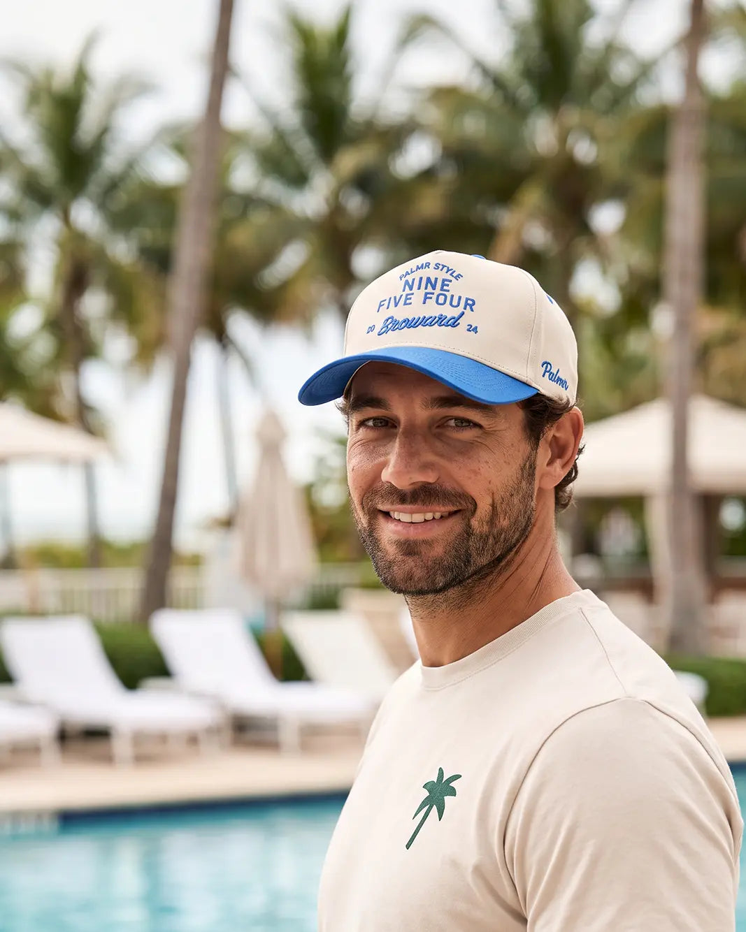 A smiling man wearing a white and blue RSV-branded cap and a cream t-shirt stands in front of a pool with palm trees in the background.