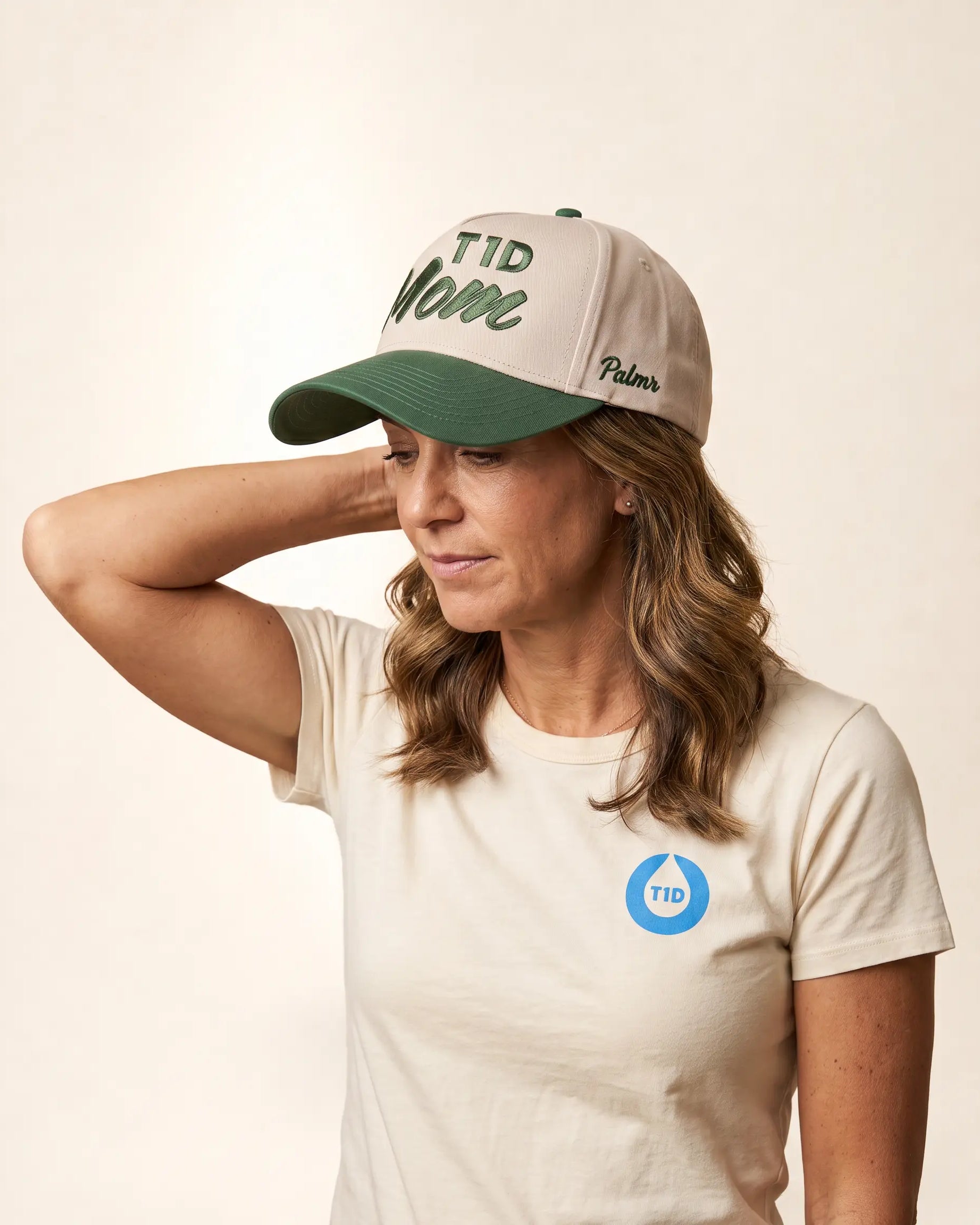 A woman wearing a beige and green baseball cap with 'TID Name' and 'Palmers' embroidered on it, paired with a matching beige T-shirt featuring a blue TID logo on the chest, set against a plain beige background.