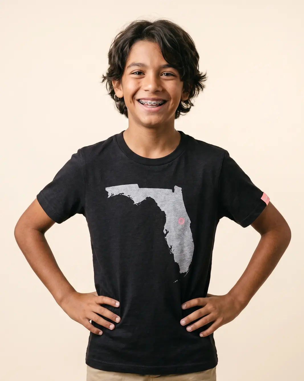 A smiling young boy with dark curly hair and braces wearing a black t-shirt with a gray Florida state graphic on the front, standing against a solid beige background.