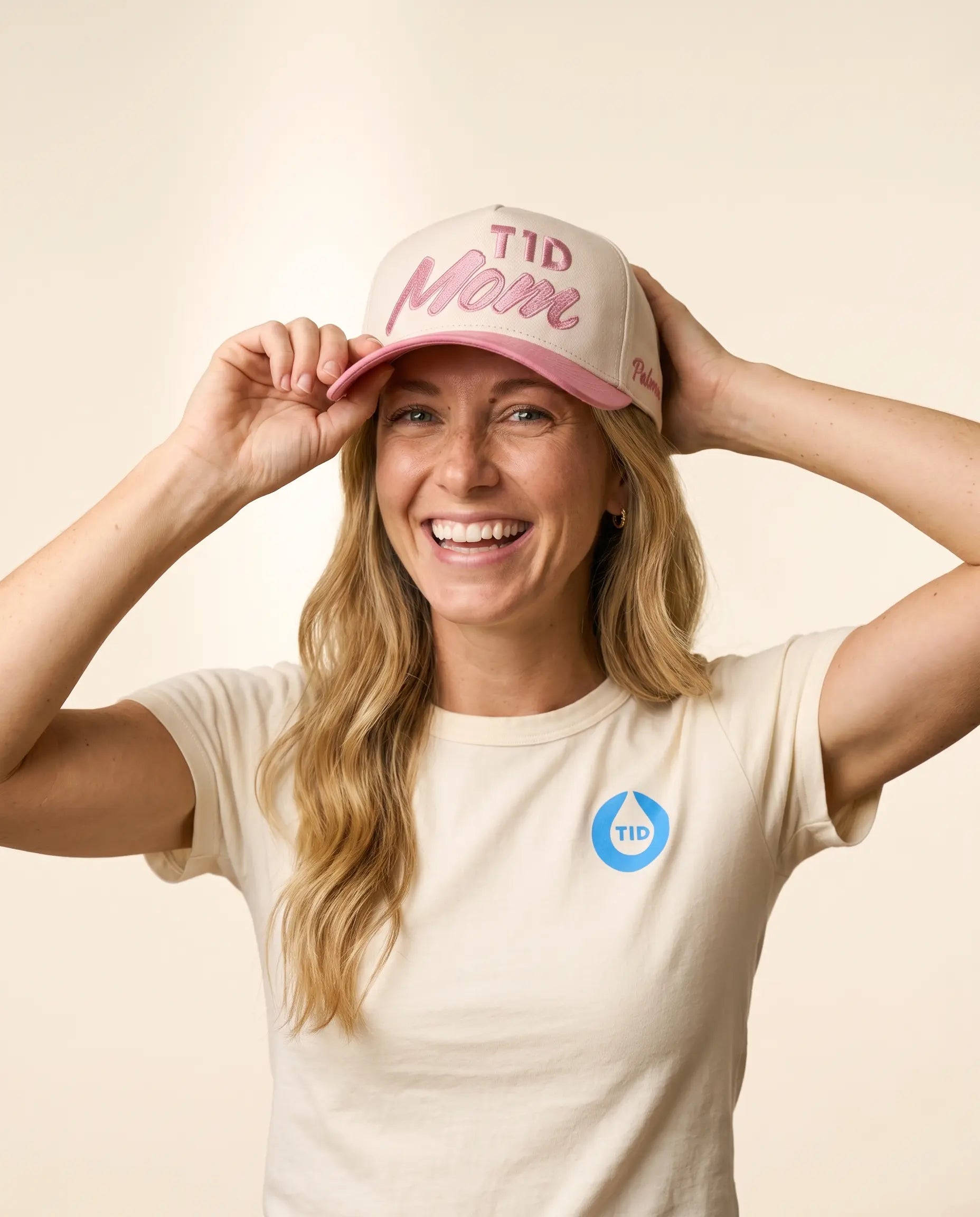 A smiling woman wearing a cream-colored T1D Mom trucker hat with pink lettering and a matching cream T-shirt with a blue T1D logo, posing against a solid cream background.