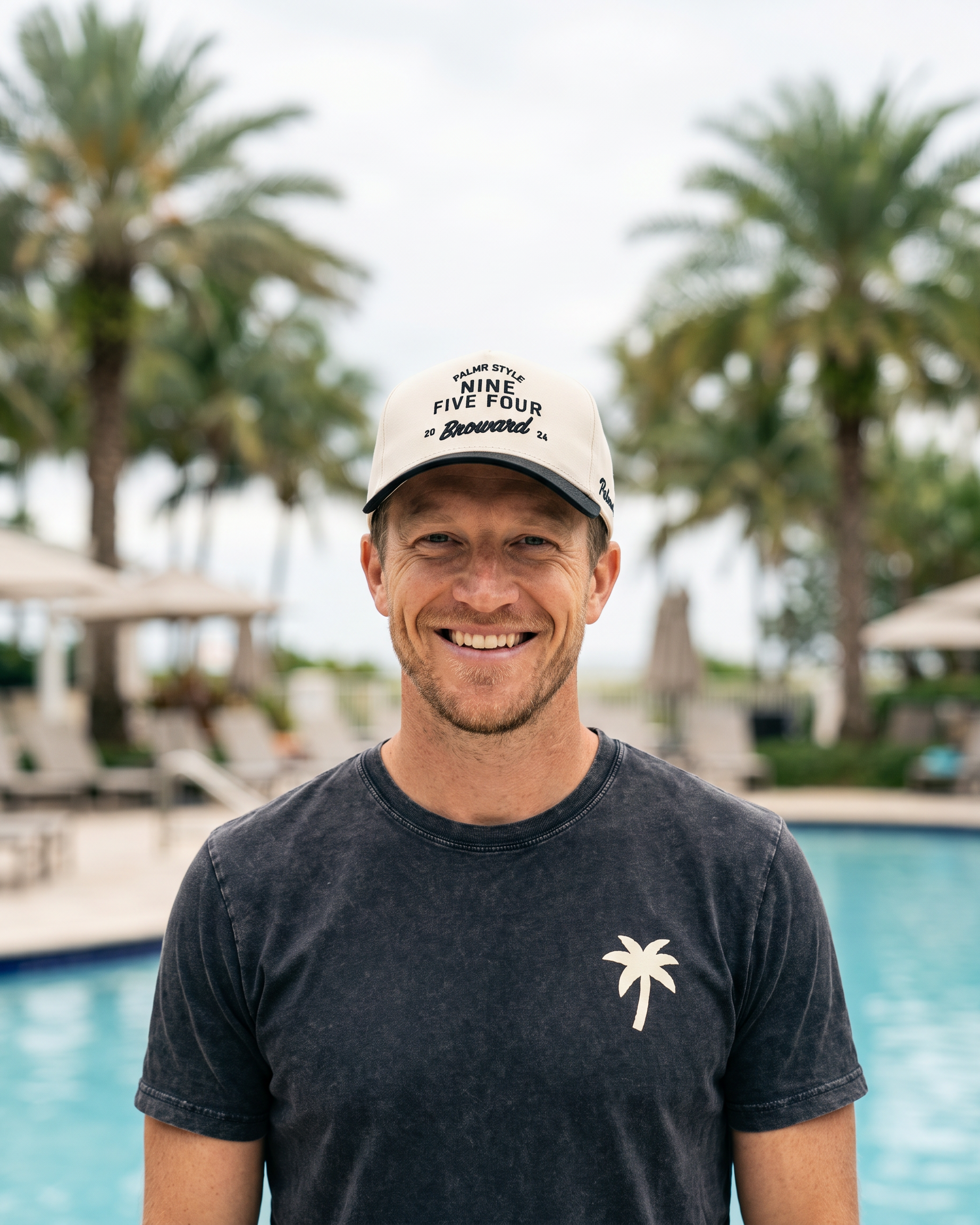 A smiling man standing in front of a swimming pool with palm trees in the background, wearing a white cap with black text and a dark charcoal t-shirt with a small white palm tree logo.