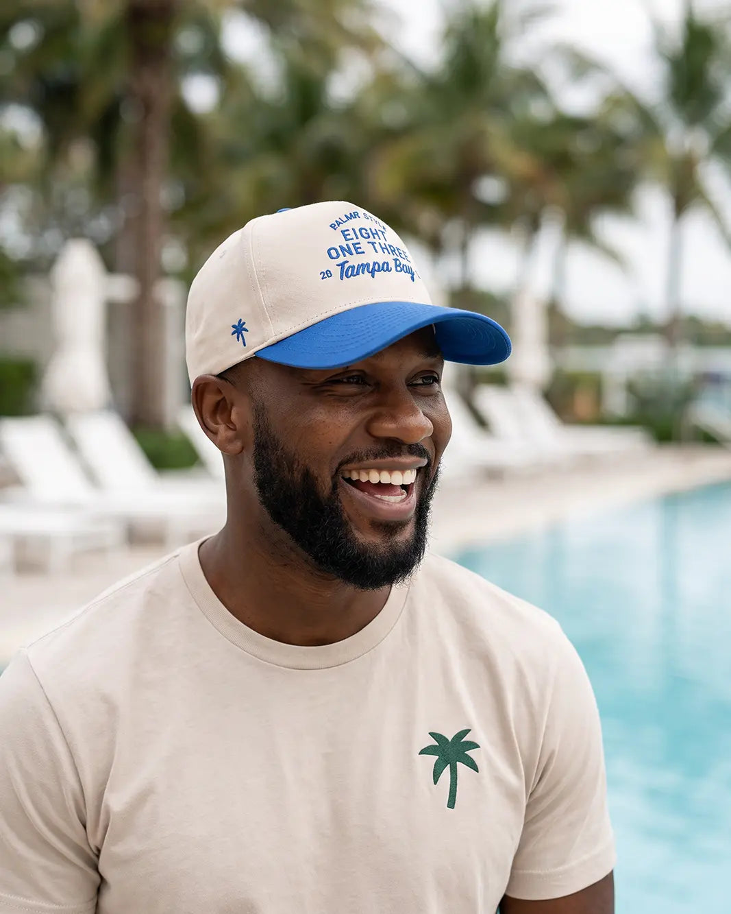 A man smiling outdoors by a swimming pool wearing a beige T-shirt with a green palm tree logo and a beige-and-blue baseball cap with blue text reading 'Welcome to One Tampa Bay'.