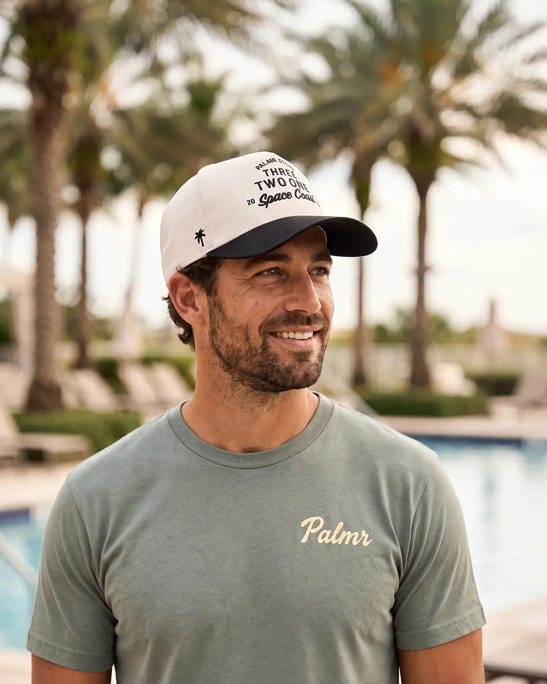 A man wearing a white and navy blue cap with 'Two Trees of Omaha' printed on it, paired with a muted sage green t-shirt featuring 'Palmr' in gold script, set against a blurred background of palm trees and a swimming pool.
