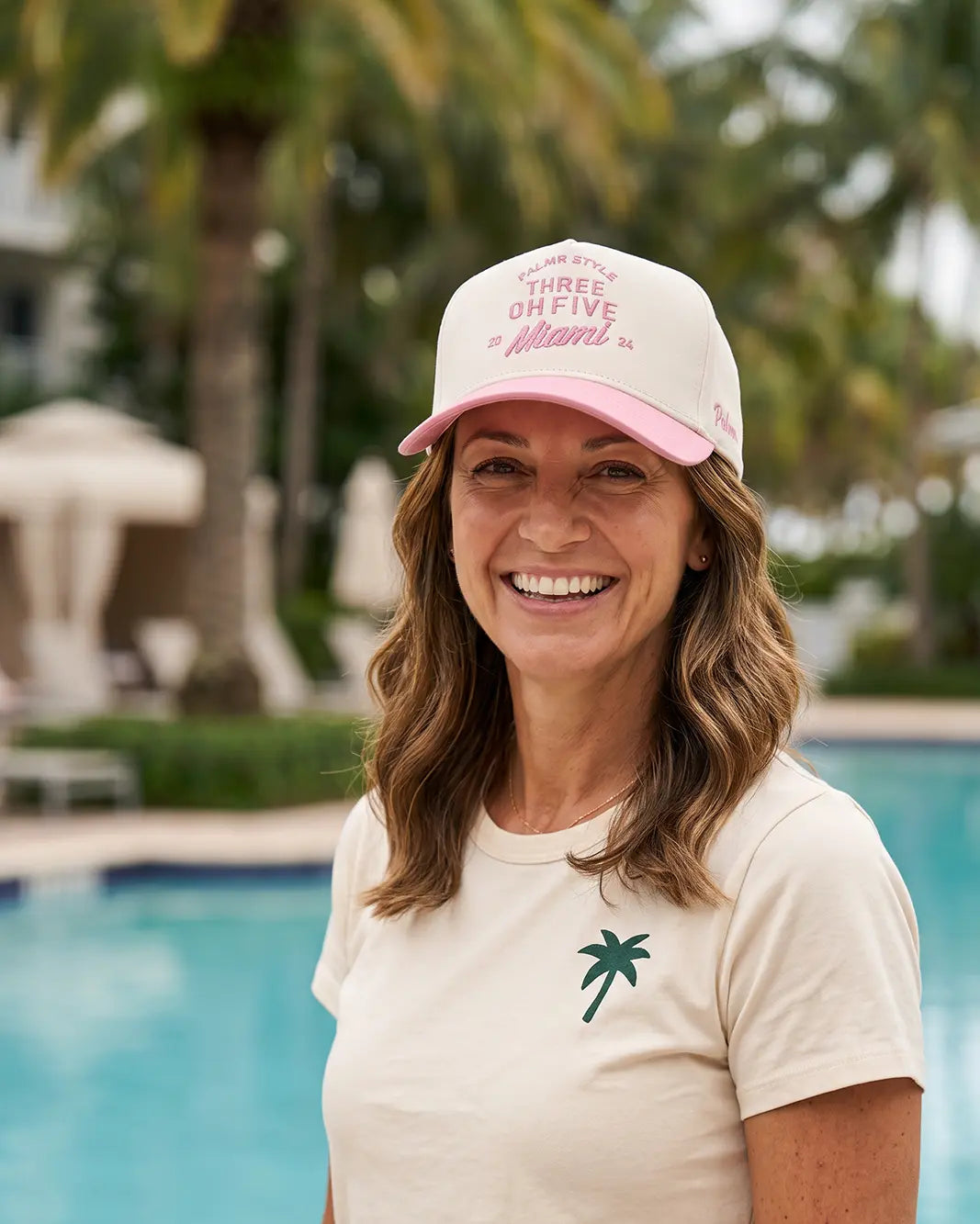 A smiling woman standing outdoors by a swimming pool wearing a cream T-shirt with a small green palm tree logo and a matching cream and pink baseball cap that reads 'Three of Five Miami'.