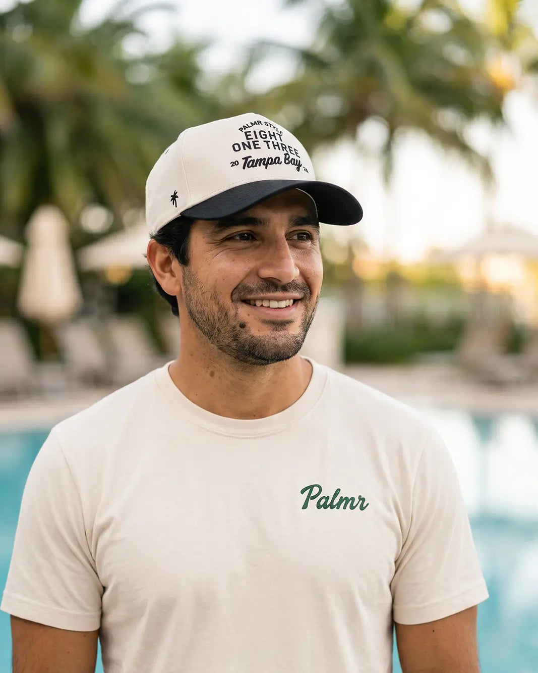 A smiling man wearing a cream-colored t-shirt with 'Palmr' printed in green on the chest and a matching cream cap with black text, set against a blurred background of a pool and palm trees.