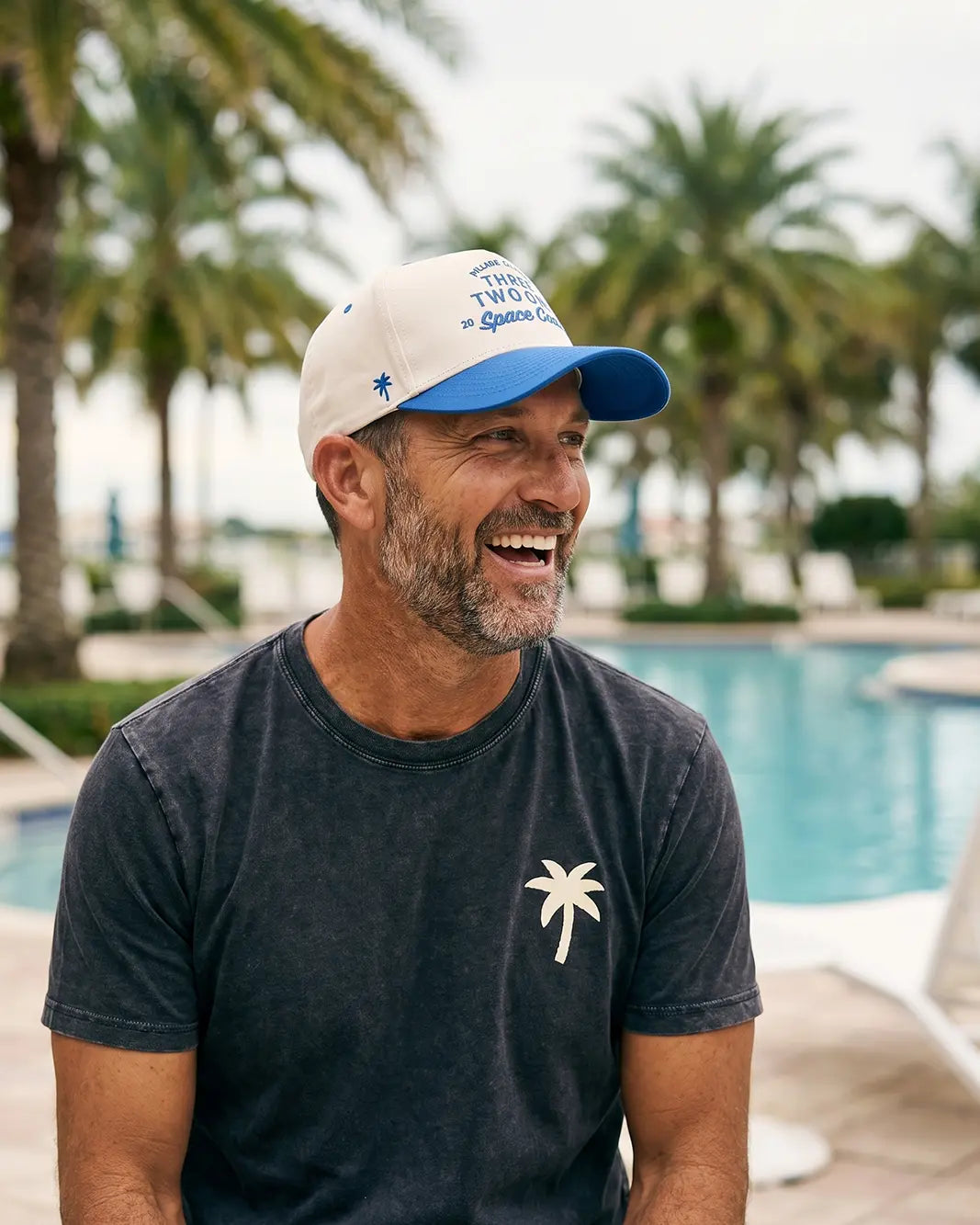 A man wearing a white and blue baseball cap with blue text and a dark gray t-shirt with a white palm tree graphic, sitting in front of a swimming pool with palm trees in the background.
