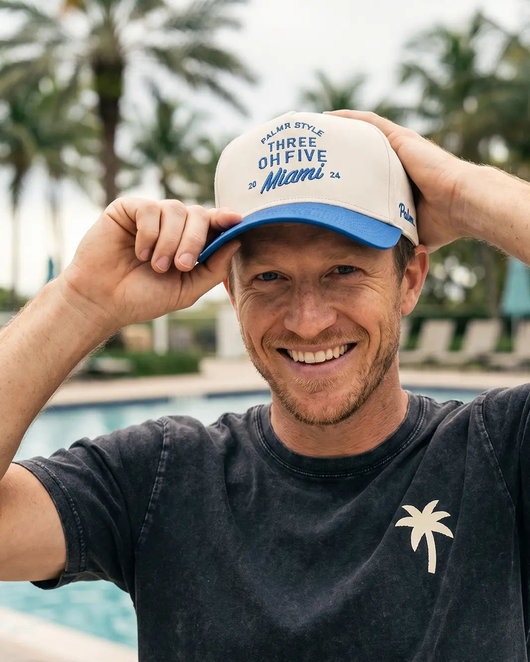 A smiling man wearing a white and blue trucker hat with 'Three Oh Five Miami' text, posing by a swimming pool with palm trees in the background.