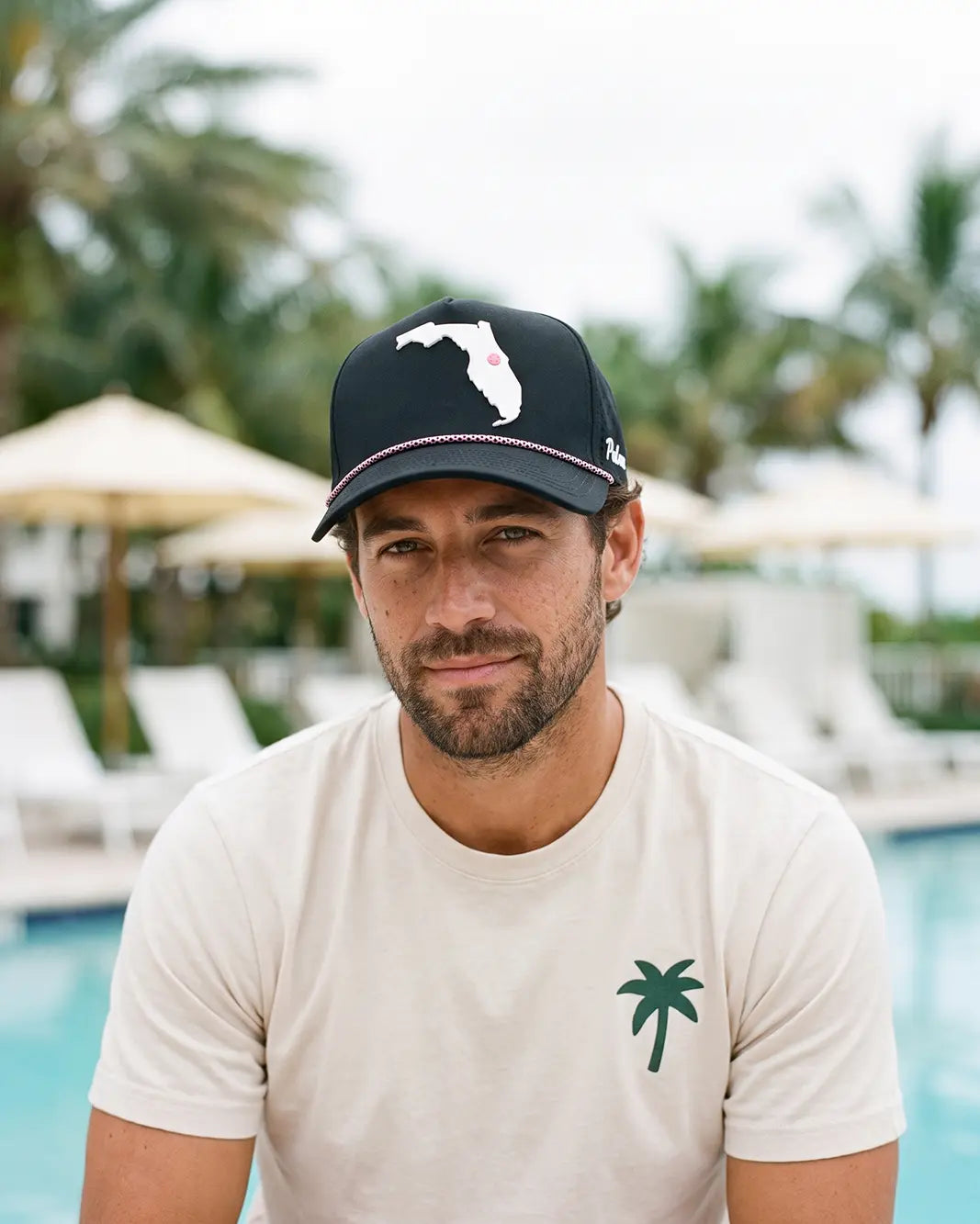 A man wearing a cream-colored t-shirt with a green palm tree logo and a black baseball cap with a white Florida state silhouette, posed in front of a pool with palm trees and lounge chairs in the background.