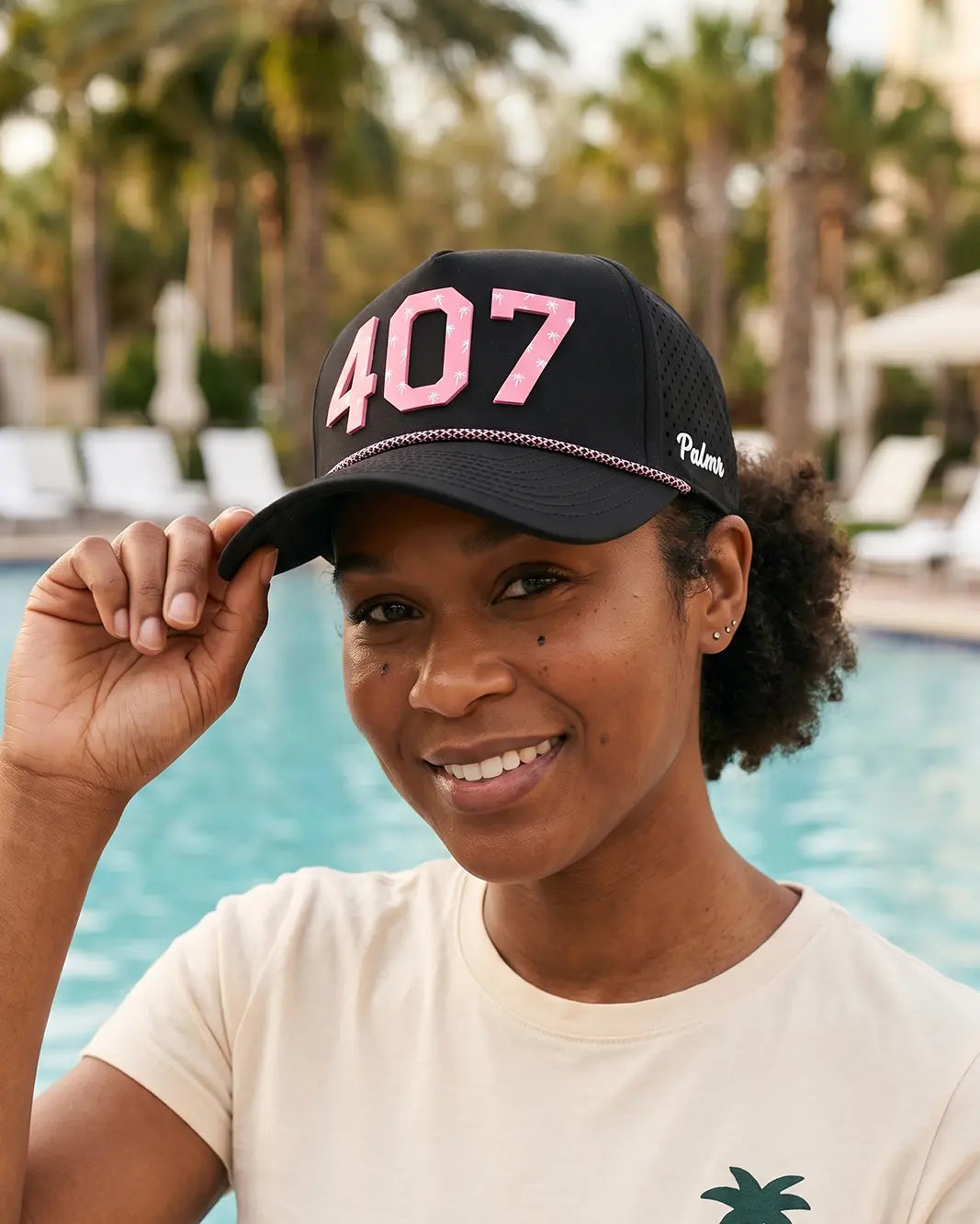 A woman wearing a black trucker hat with pink '407' embroidery and a small white 'Palmr' logo, posing in front of a swimming pool with palm trees in the background.
