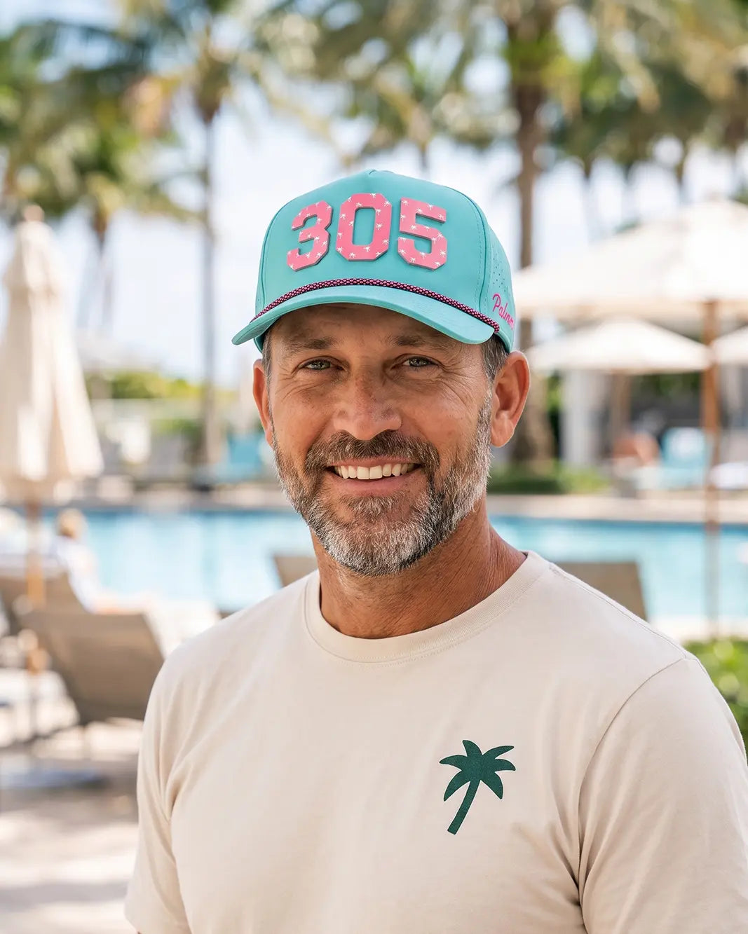 A smiling man wearing a turquoise baseball cap with pink '305' lettering and a beige T-shirt with a green palm tree logo, photographed outdoors in front of a blurred resort pool area with palm trees.