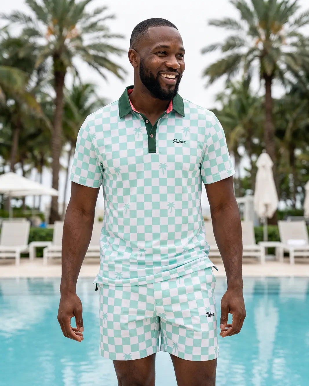 A man wearing a mint green and white checkered polo shirt and matching shorts stands in front of a swimming pool with palm trees in the background. The brand name 'Palmers' is visible on the chest of the polo shirt.