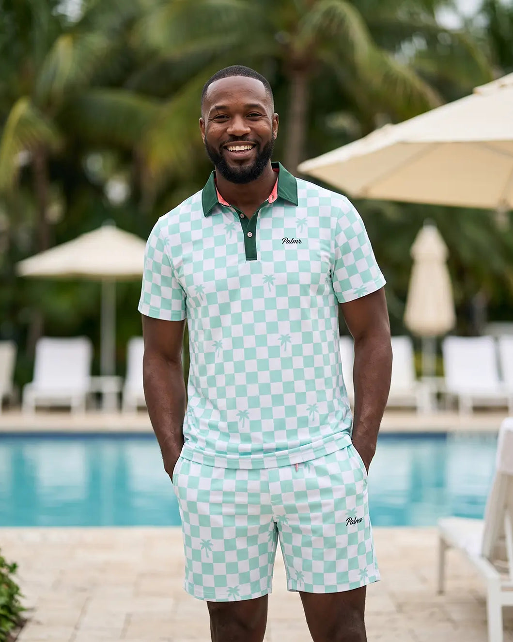 A man wearing a light green and white checkered polo shirt and matching shorts stands in front of a swimming pool with palm trees and umbrellas in the background. The brand name 'Paisley' is visible on the chest of the polo shirt.