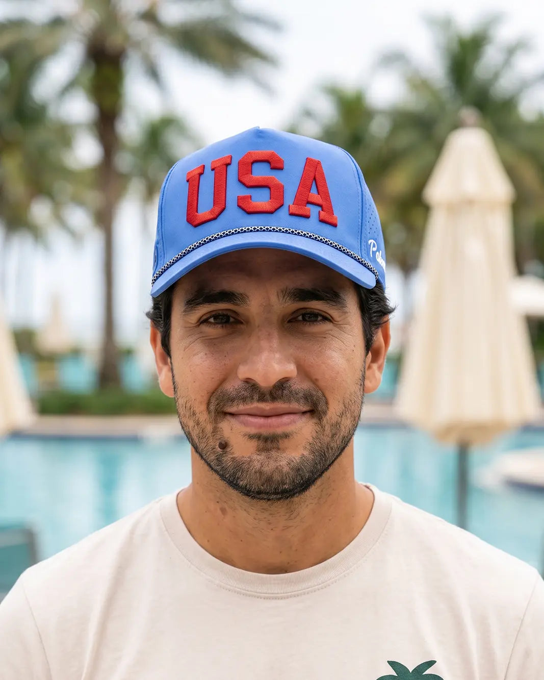 A man wearing a blue baseball cap with 'USA' in red letters, photographed outdoors in front of a pool area with palm trees and closed umbrellas.