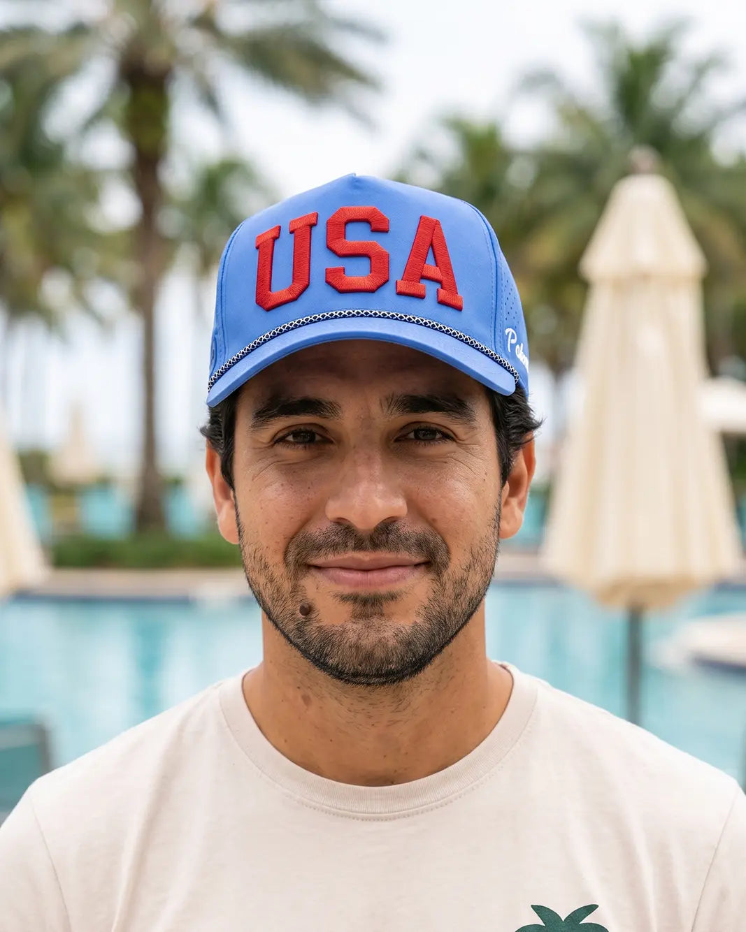 A man wearing a blue baseball cap with 'USA' in red letters, photographed outdoors in front of a pool area with palm trees and closed umbrellas.