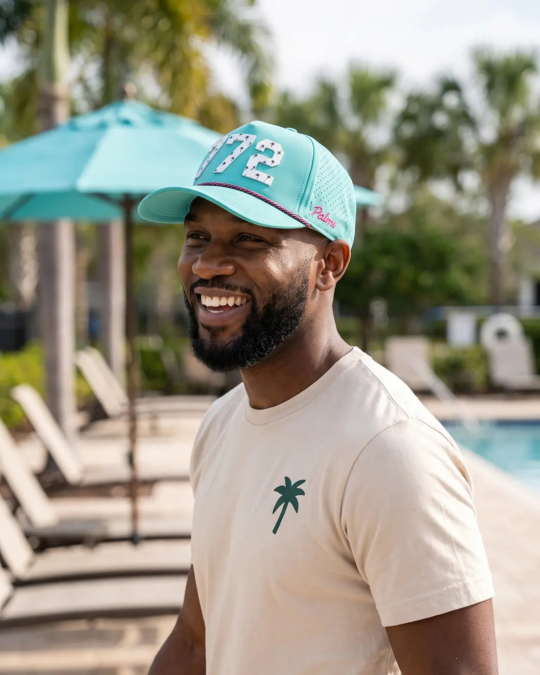 The 772 Signature Hat (Teal) worn on-model poolside, smiling, three-quarter view: white 772 with palm print on a teal crown, pink rope, pink Palmr script.
