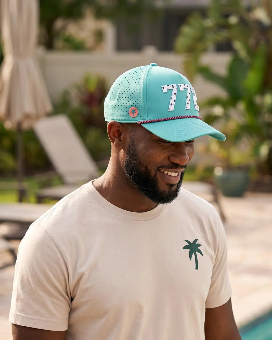The 772 Signature Hat (Teal) worn on-model by the pool, head tilted down, side view: perforated panel, pink palm badge, white 772 and pink rope.