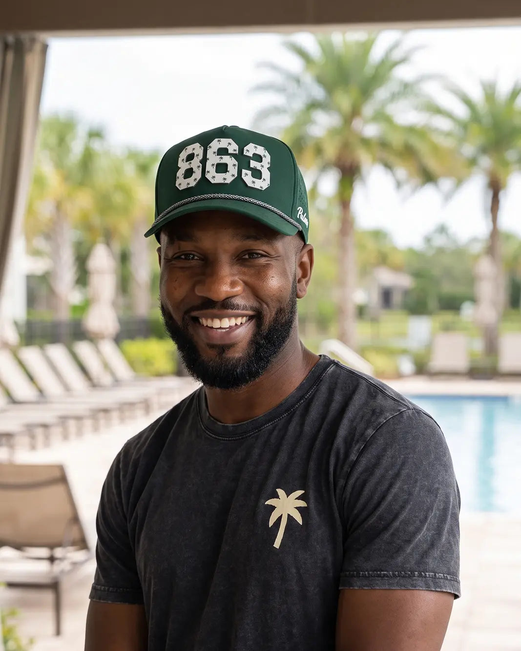The 863 Signature Hat (Green) worn on-model poolside, smiling, front view: white 863 with palm print on a dark green crown, pink rope.