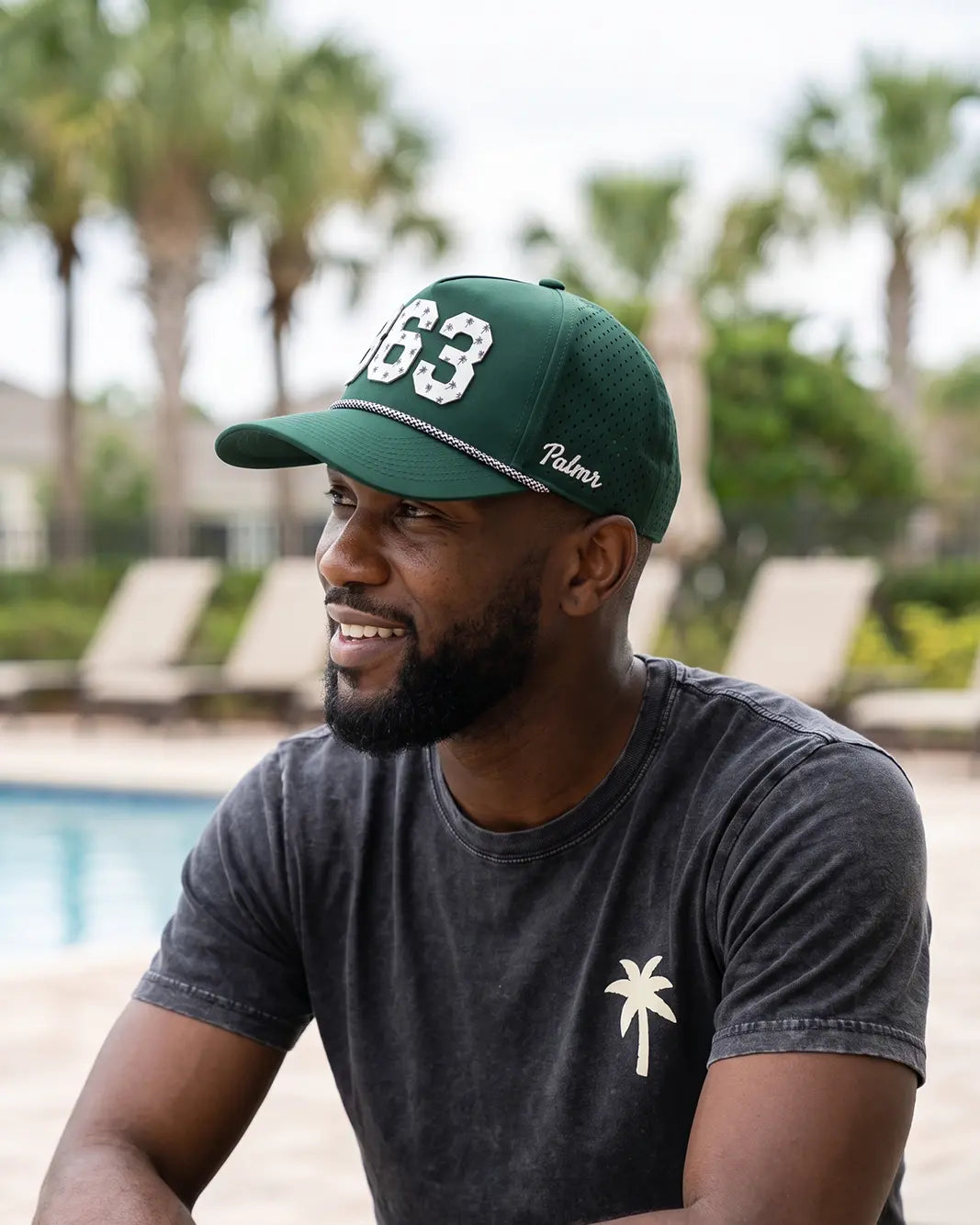 The 863 Signature Hat (Green) worn on-model, seated by the pool, three-quarter view: white 863 on a dark green crown, perforated panel, white Palmr script.