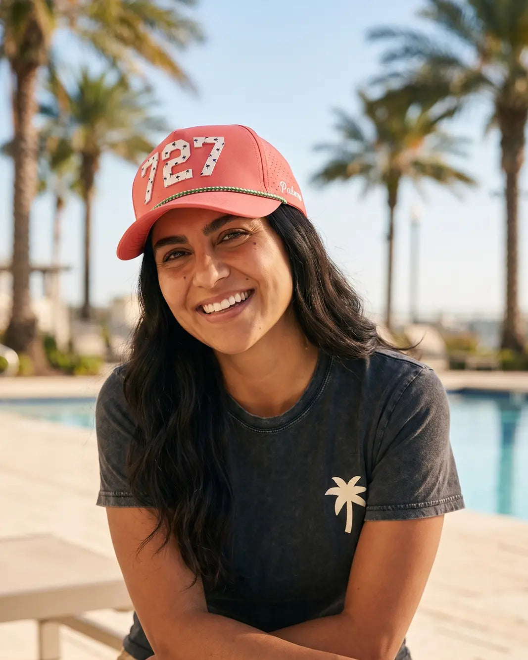 The 727 Signature Hat (Pink) worn on-model, seated poolside, front view: white 727 with palm print on a pink crown, green rope, Palmr script.