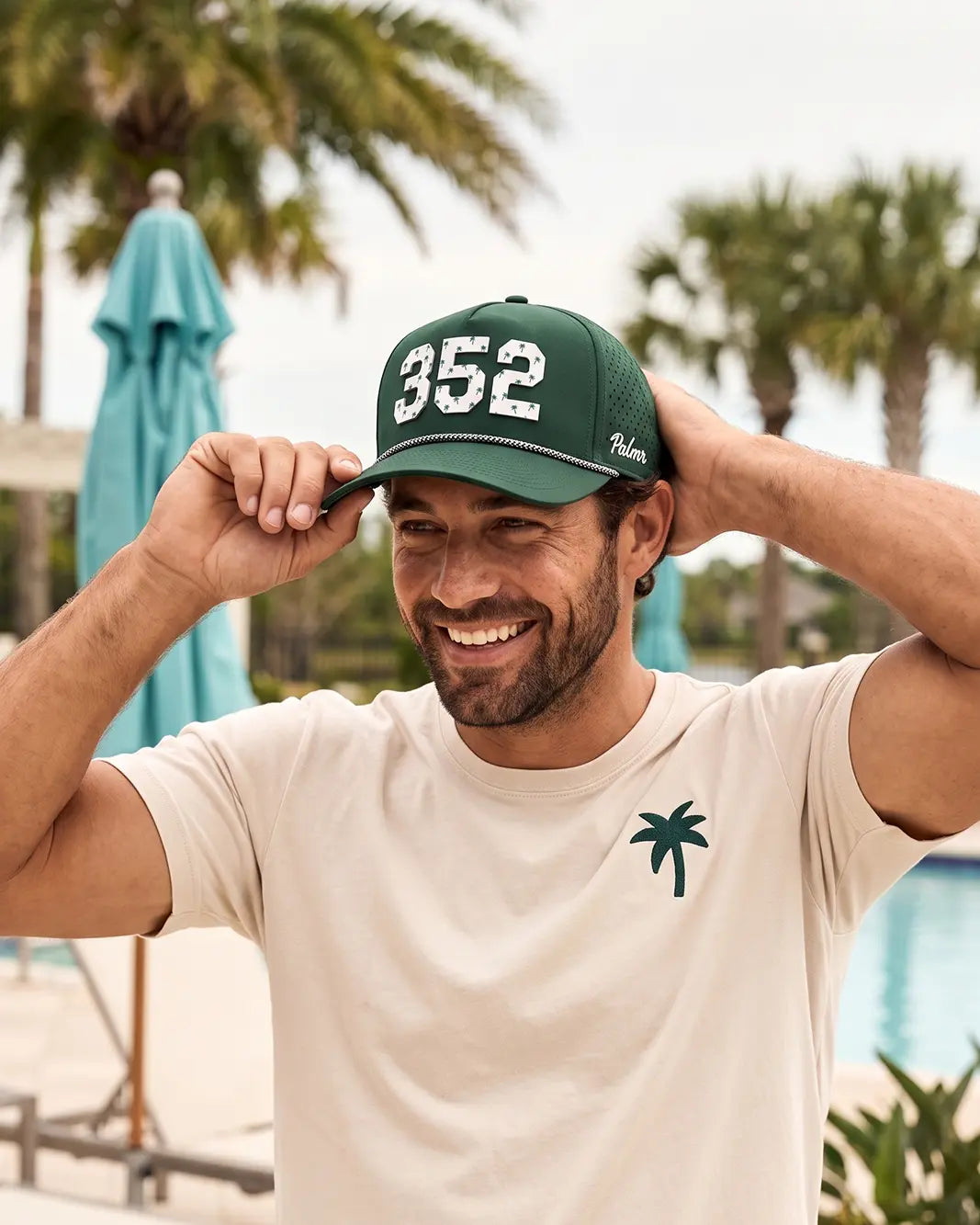 The 352 Signature Hat (Green) worn on-model poolside, both hands adjusting the hat: white 352 on a dark green crown, white Palmr script.