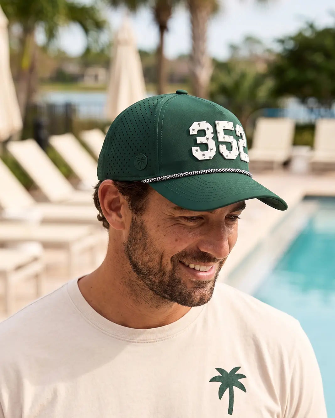 The 352 Signature Hat (Green) worn on-model by the pool, head tilted down, three-quarter view: white 352 with palm print on a dark green crown, perforated panel.