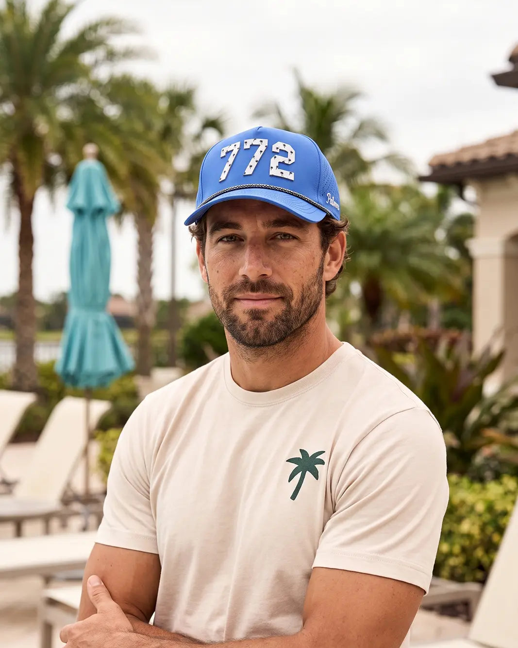 The 772 Signature Hat (Blue) worn on-model on a pool deck, straight-on view: white 772 with palm print on a blue crown, black and white rope.