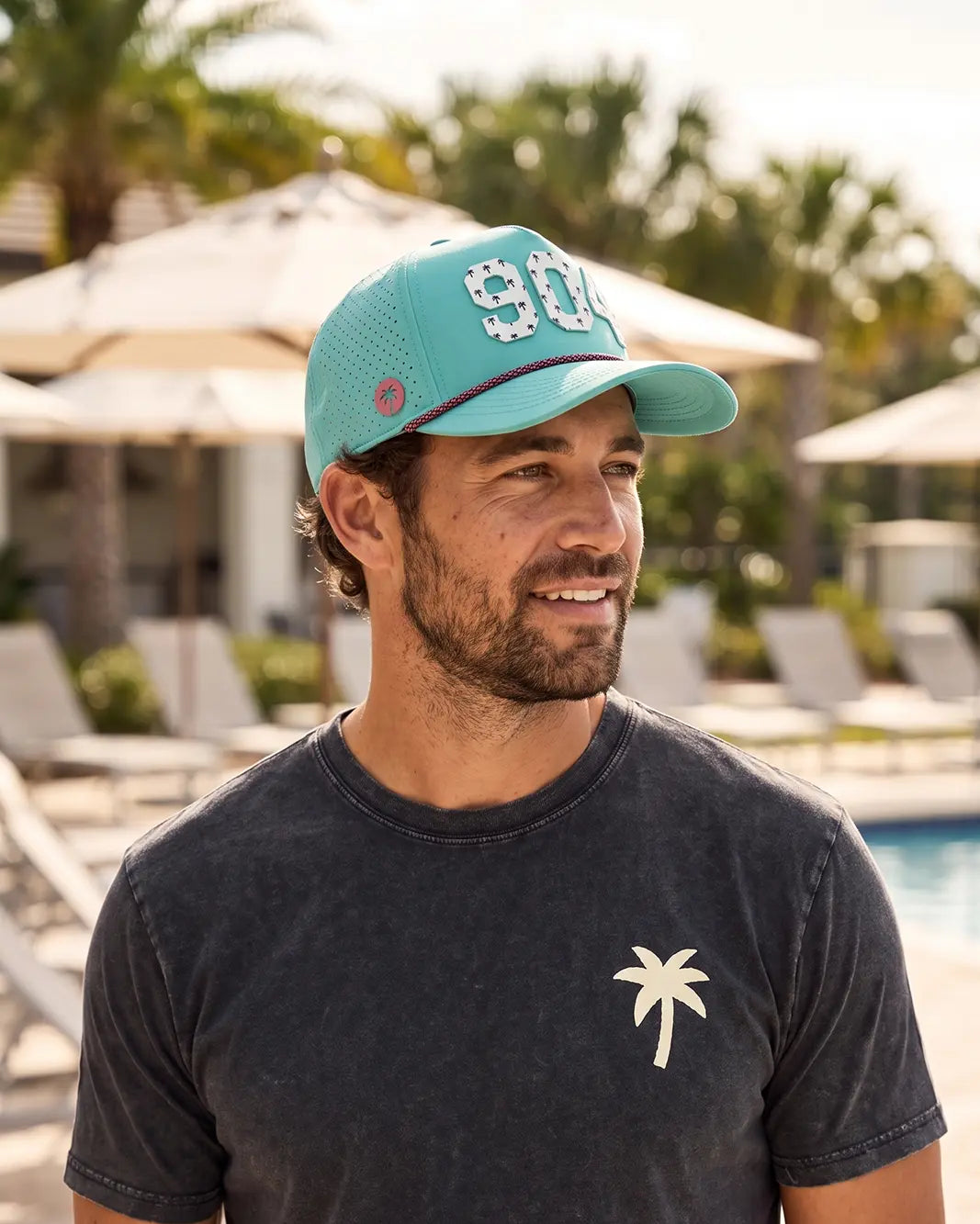 The 904 Signature Hat (Teal) worn on-model poolside, side profile: perforated panel, pink palm badge, white 904 and pink rope on a teal crown.