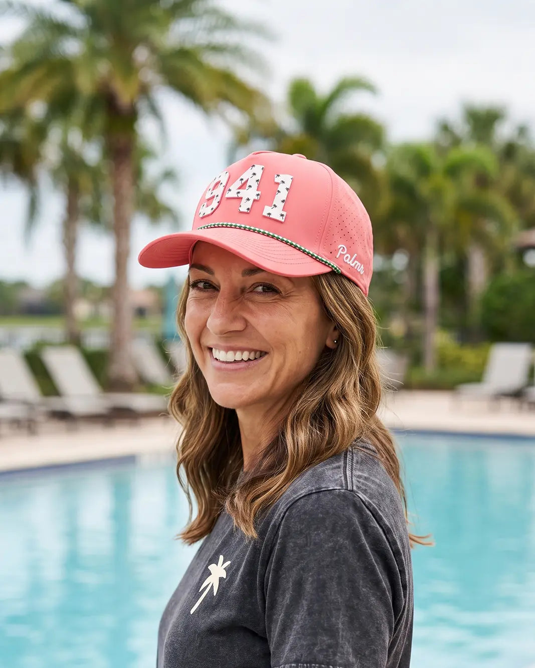 The 941 Signature Hat (Pink) worn on-model poolside, looking back over the shoulder: white 941 with palm print on a pink crown, green rope, Palmr script.