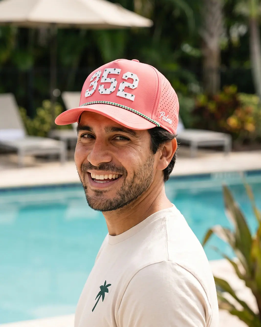 The 352 Signature Hat (Pink) worn on-model poolside, looking back over the shoulder: white 352 on a pink crown, green rope, Palmr script.