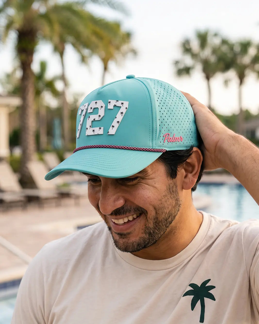 The 727 Signature Hat (Teal) worn on-model poolside, hand on the back of the hat, head down: white 727 on a teal crown, pink rope, pink Palmr script.