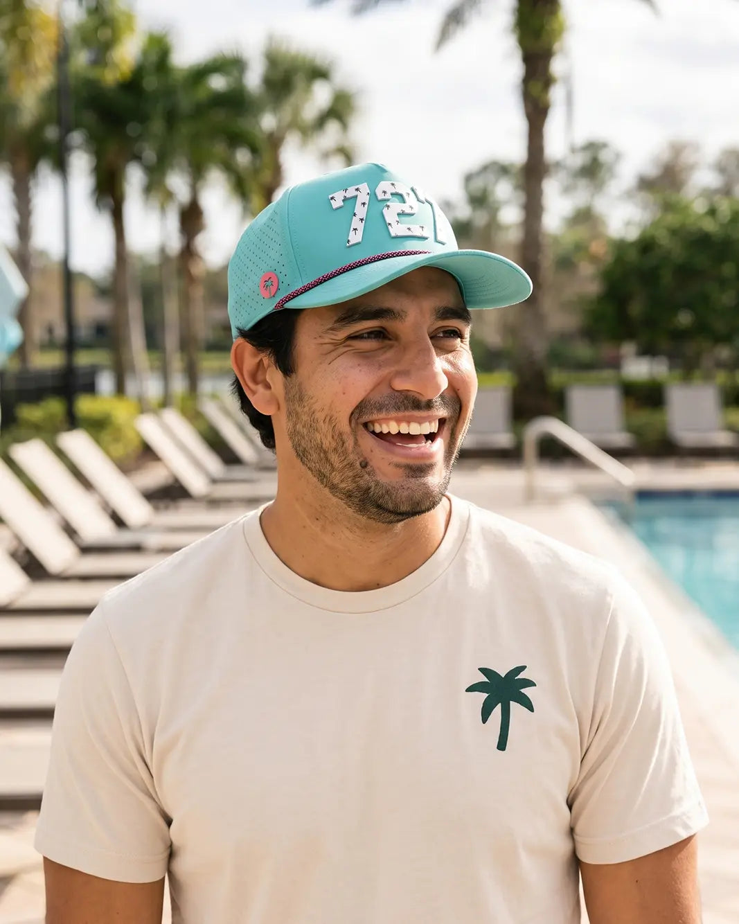 The 727 Signature Hat (Teal) worn on-model by the pool, laughing, three-quarter view: white 727 with palm print on a teal crown, pink rope.