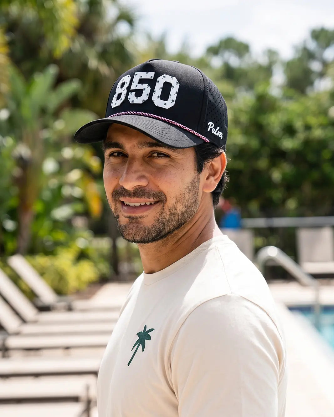 The 850 Signature Hat (Black) worn on-model poolside, looking back over the shoulder: white 850 with palm print on a black crown, pink rope, Palmr script.