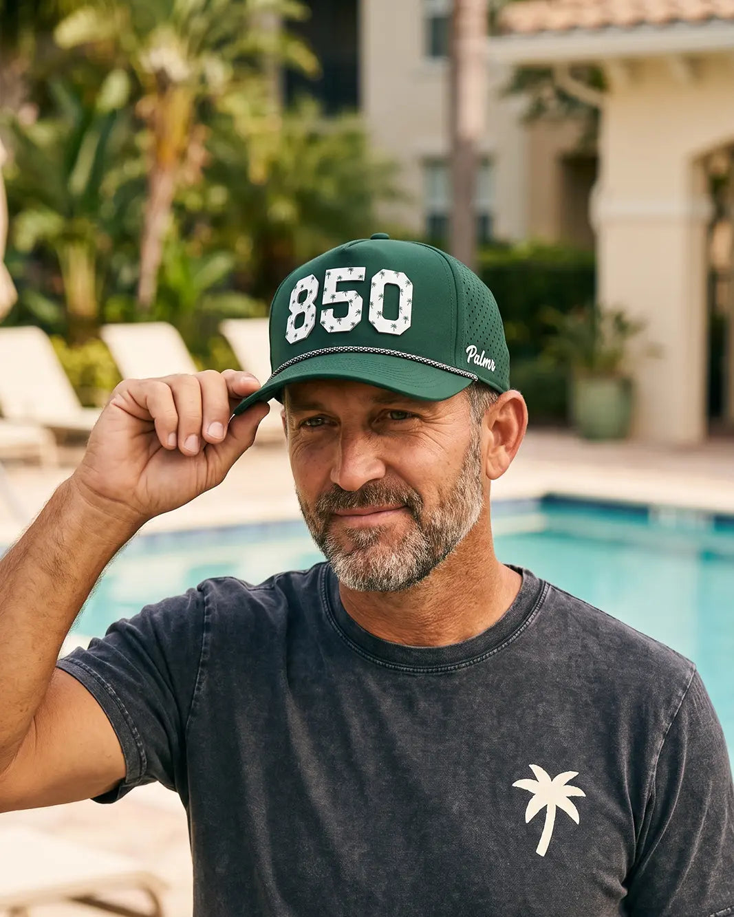 The 850 Signature Hat (Green) worn on-model poolside, hand on the brim: white 850 with palm print on a dark green crown, pink rope, Palmr script.