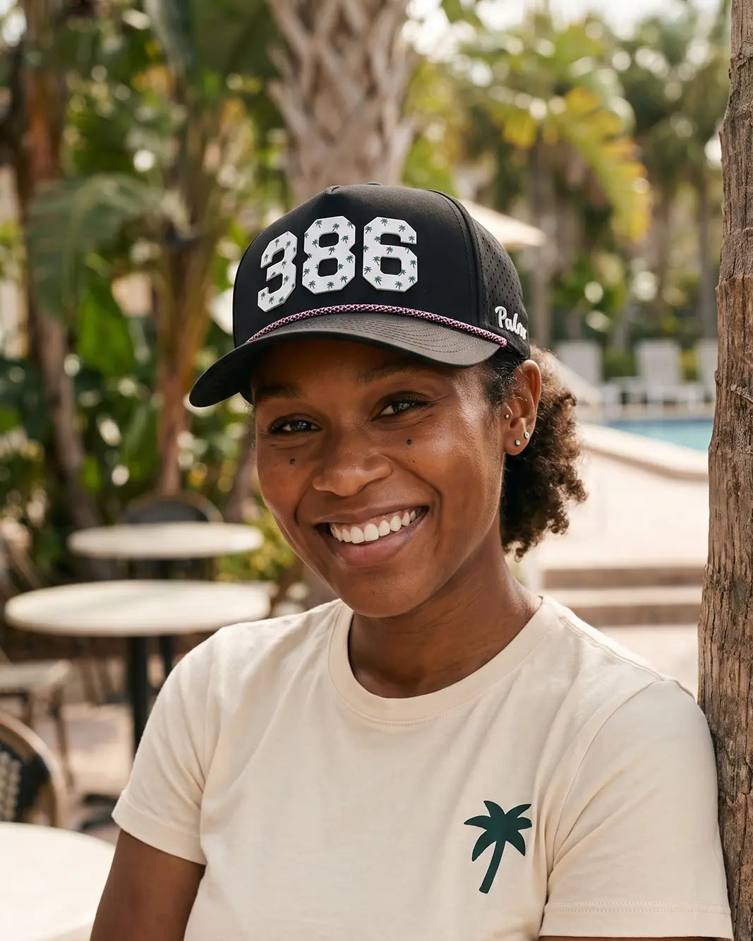The 386 Signature Hat (Black) worn on-model poolside, front three-quarter view: white 386 with palm print on a black crown, pink rope, Palmr script.
