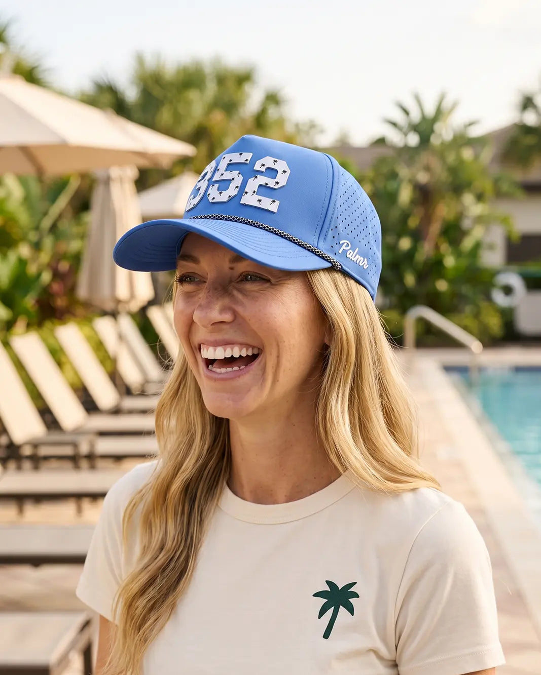 The 352 Signature Hat (Blue) worn on-model poolside, laughing, three-quarter view: white 352 and Palmr script on a blue crown.