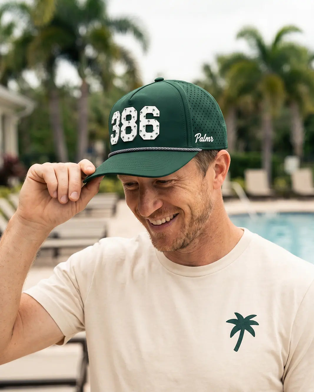 The 386 Signature Hat (Green) worn on-model poolside, hand on the brim: white 386 with palm print on a dark green crown, pink rope, Palmr script.