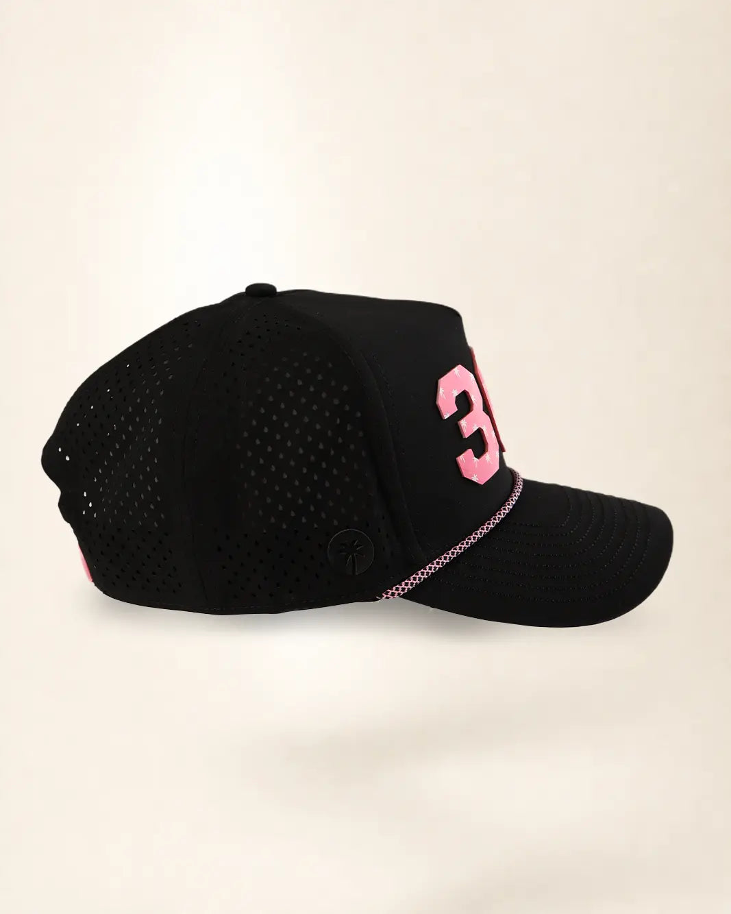 A black baseball cap with a perforated front panel, featuring a pink number '3' on the side and a pink-and-white dotted line detail along the edge of the brim, set against a plain light beige background.